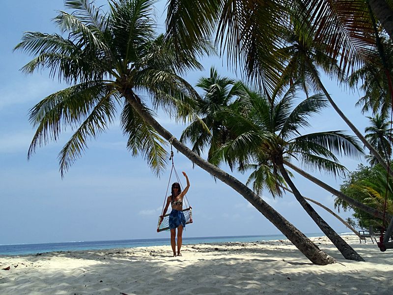 Woman standing on a swing beneath leaning palm trees on a white sand beach in Maalhos, Maldives.